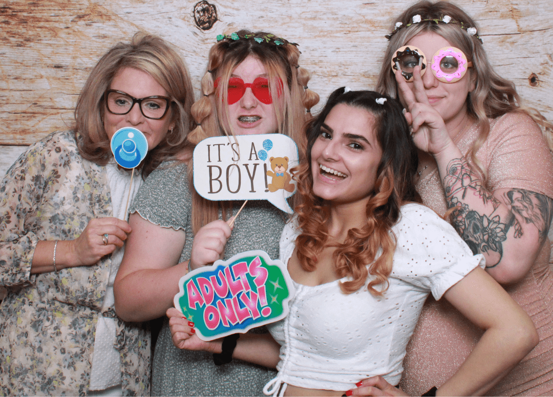 A group of women enjoying a fun moment at a baby shower, posing with props in a photo booth.