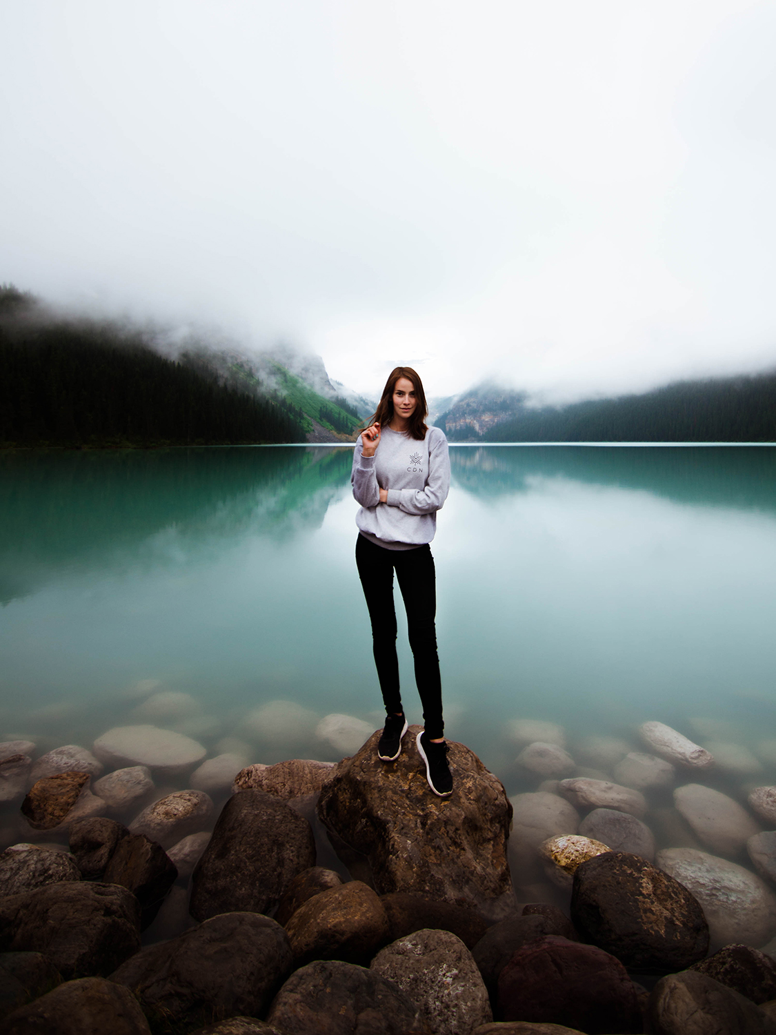 Female subject stands on rock on Lake Louise on cloudy sky day. The water is beautifully clear