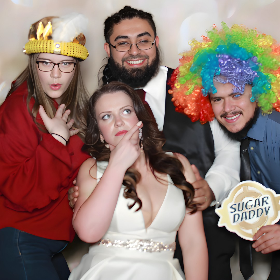 Group of friends posing with fun props and a 'Sugar Daddy' sign in a photo booth.