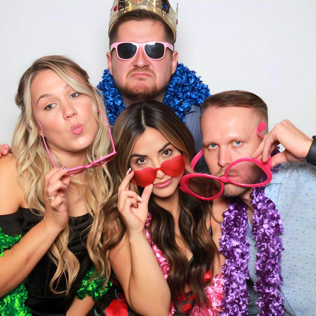 Group of four friends posing with fun props in a photo booth, wearing sunglasses, a crown, and boas.